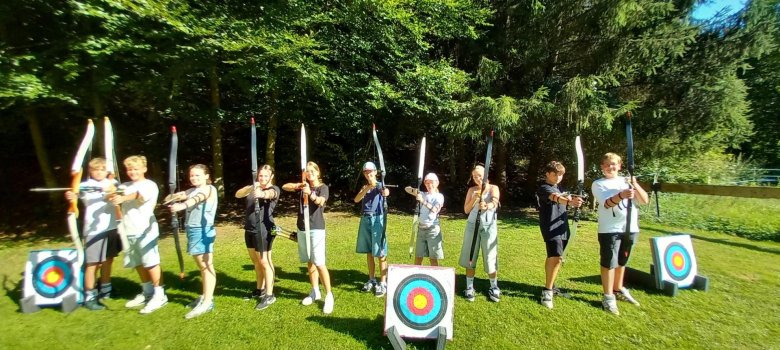 Bogenschießen Gruppenbild aller Workshop-Teilnehmer mit Pfeil und Bogen auf der Hand, den Pfeil schussbereit auf die Kamera gerichtet. Im Hintergrund eine grüne Wiese mit den Zielbögen.