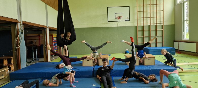 Akrobatik Gruppenbild der Workshopteilnehmer, die verschiedene akrobatische Übungen, wir Brücken, Luftspagat auf den Turnmatten in einer Turnhalle präsentieren.