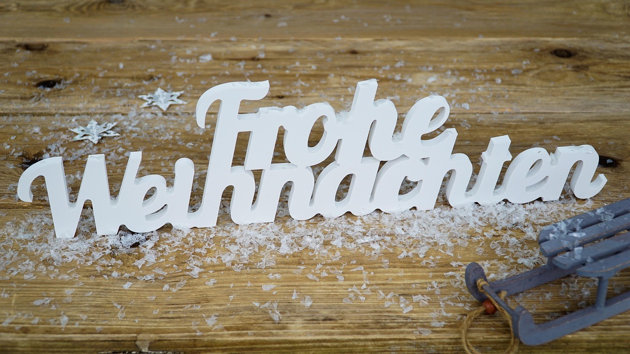 Der Schriftzug "Frohe Weihnachten" auf einer Holzunterlage mit Kunstschnee bedeckt und einem kleinen Dekoschlitten an der Seite.
