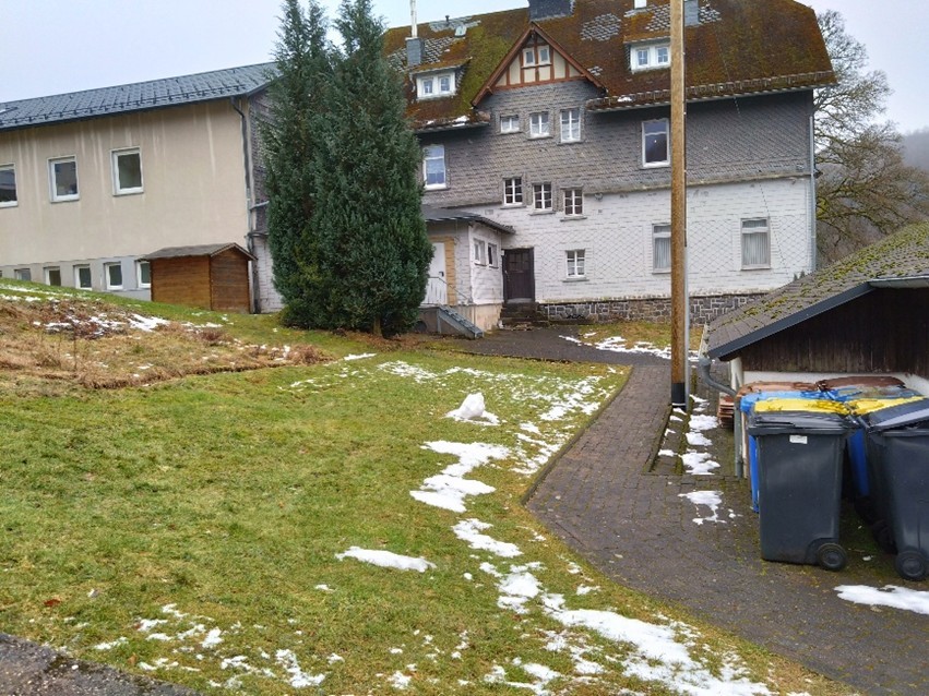 Zugangsweg zu den Mietwohnungen des Dorfgemeinschaftshauses, mit Wiese und Mülltonnen im Vordergrund.