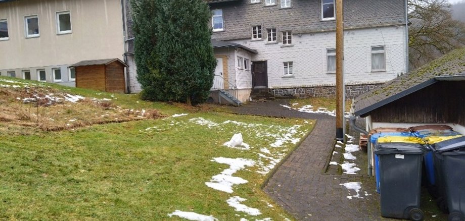 Zugangsweg zu den Mietwohnungen des Dorfgemeinschaftshauses, mit Wiese und Mülltonnen im Vordergrund.