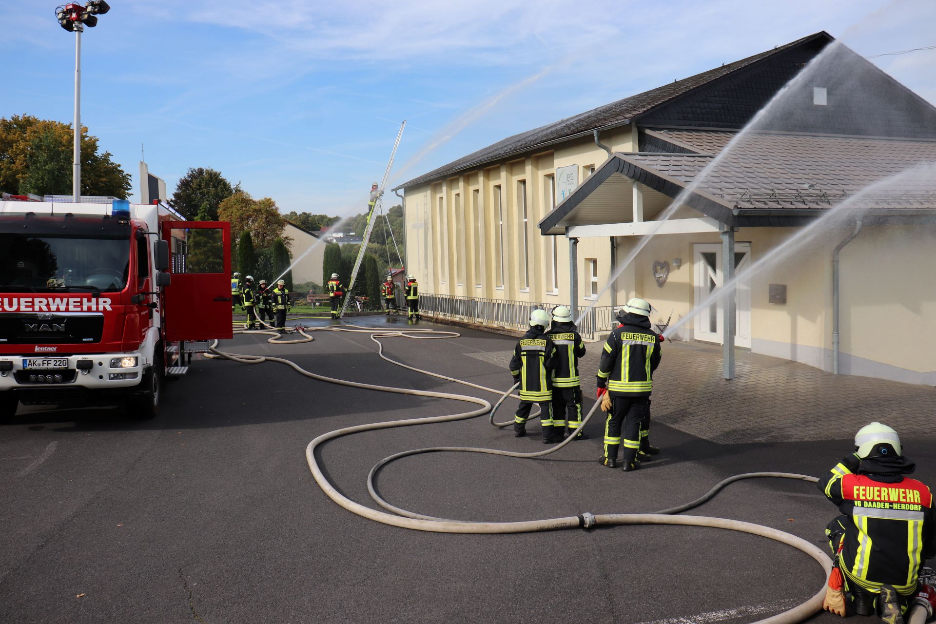 Hier sehen Sie ein Feuerwehrauto sowie mehrere Feuerwehrleute beim Löschen eines gelben Gebäudes.