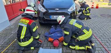 Hier sehen Sie vier Feuerwehrleute in Uniform und Ausrüstung beim Versorgen einer Übungspuppe, die halb unter einem Auto liegt.