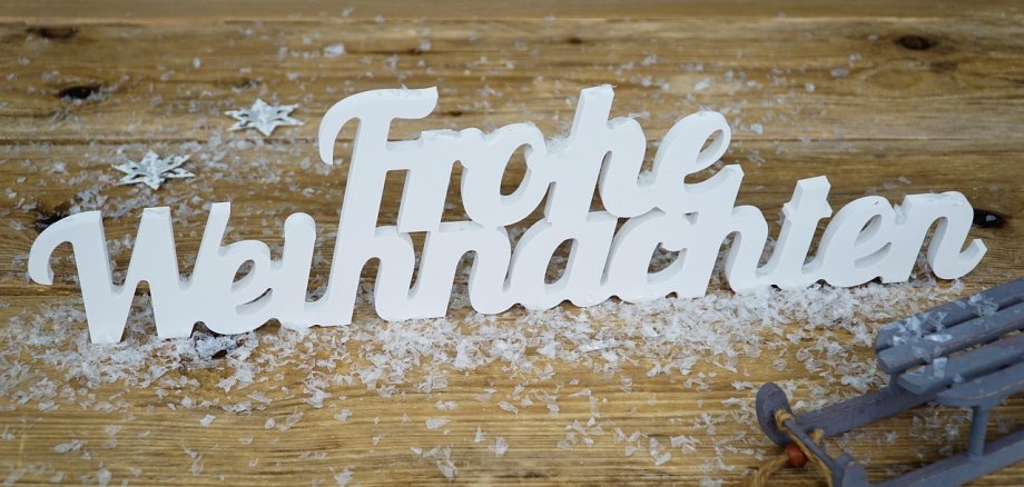 Der Schriftzug "Frohe Weihnachten" auf einer Holzunterlage mit Kunstschnee bedeckt und einem kleinen Dekoschlitten an der Seite.