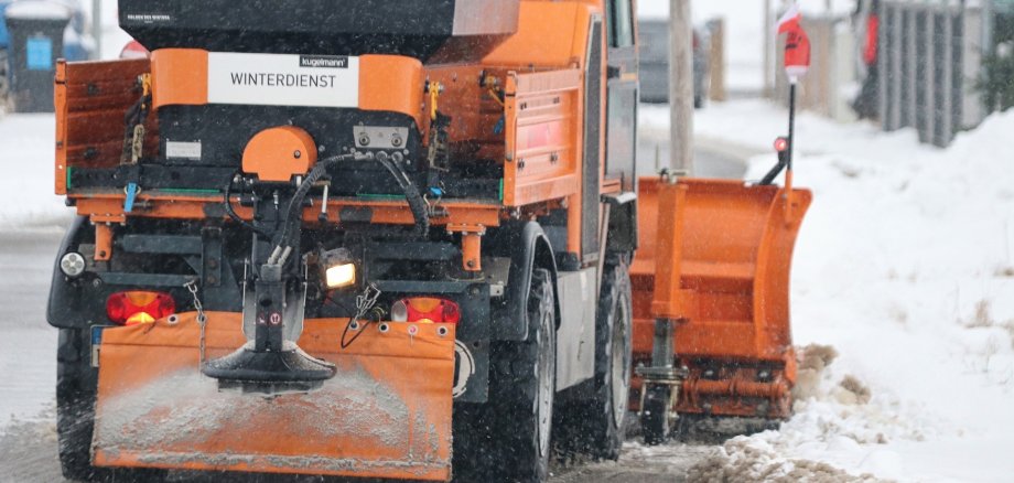 Ein Schneepflug von hinten der eine verschneite Straße räumt und Salz streut.