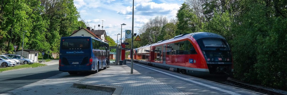 Hier sehen Sie eine Zug und einen Bus die parallel an zwei Haltstellen halten.
