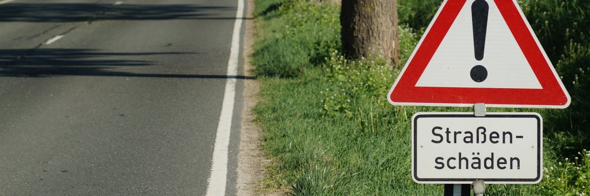 Hier sehen Sie das Verkehrsschild "Allgemeine Gefahrenstelle" mit dem Hinweis "Straßenschäden" und im Hintergrund eine Landstraße.